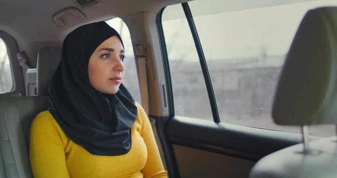 Young muslim woman in hijab sitting in car on passenger rear seat. Muslim woman day dreaming in back seat. A young woman in a hijab looks out the window.