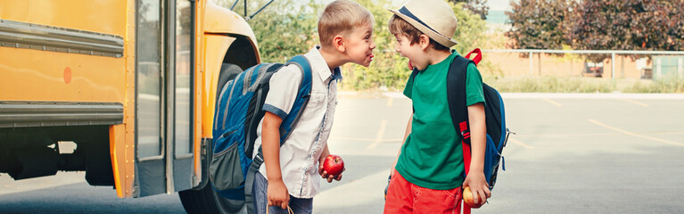 Two funny happy Caucasian boys kids showing tongues each other near yellow bus on 1 September day. Education back to school. Children learn and study. Web banner header for a website.