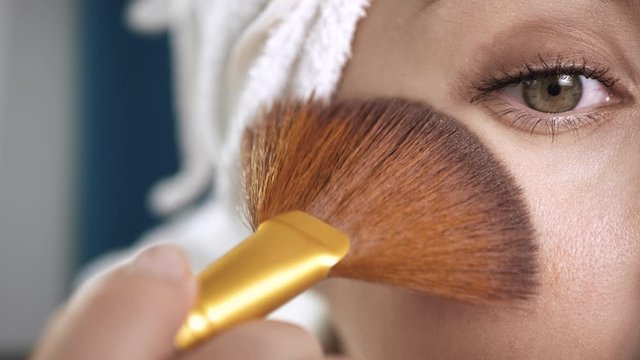 Girl applying on face shine for skin. Macro shot of attractive woman with towel puts highlighter or bronzer on skin. Make-up artist, beauty, body care, preparation of date or celebration concept