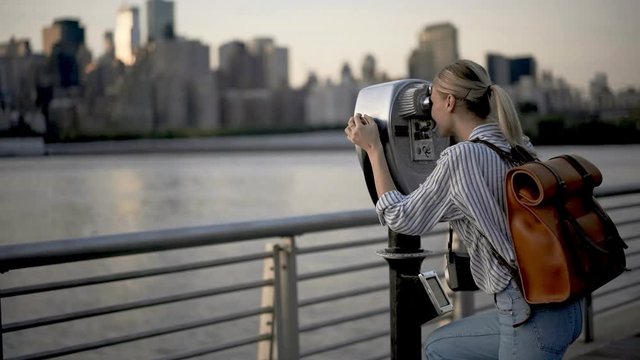 Smiling hipster girl with touristic backpack enjoying evening promenade at Hudson river using optical tower viewer for looking at scenic Manhattan island, Caucasian woman photographing cityscape 
