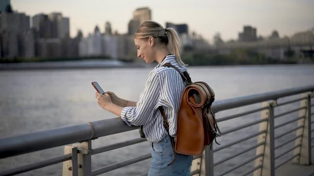 Millennial female tourist using cellphone for online networking during American vacations, young woman photographing Manhattan island while spending leisure near Hudson river
