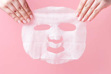 Female hands hold white fabric mask on pink background.