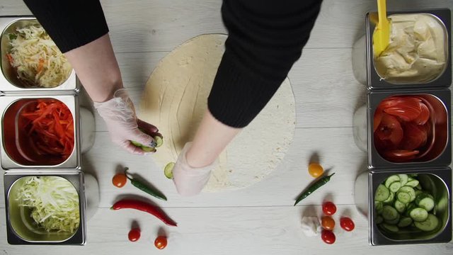 Top view of chef hands in white gloves puts cucumbers on doner kebab shawarma in pita or lavash. Shawarma with chicken cutlet, pappers, cheese and vegetable