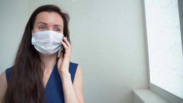 Attractive smiling brunette woman in blue dress wear protective medical face mask, talk on mobile phone about her recover near white hospital window. Healing concept in pandemic by COVID-19 threat.