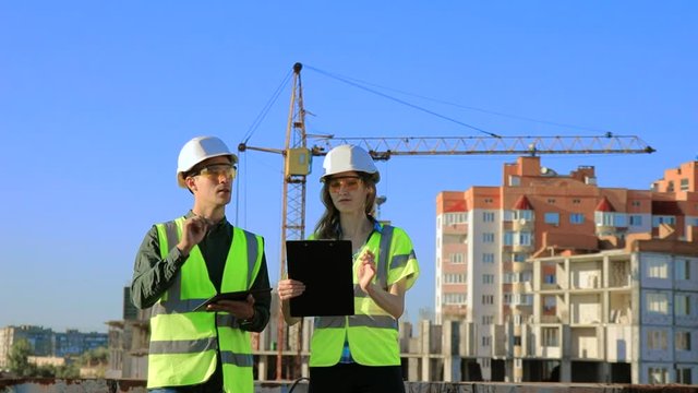 Young engineers architects guy and girl in protective helmet and vest about the project on a tablet and notebook against the background of a construction crane.