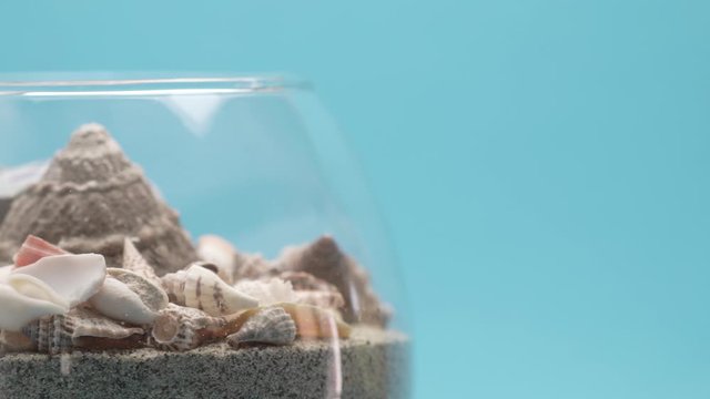 Small shells from the Greek sea lined on sand in a glass vase spin in a circle on a turquoise blue background