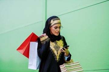 Stunning arab woman in traditional wear walking outdoors with shopping bags in hands and typing or reading message on smart phone. Millennial generation.