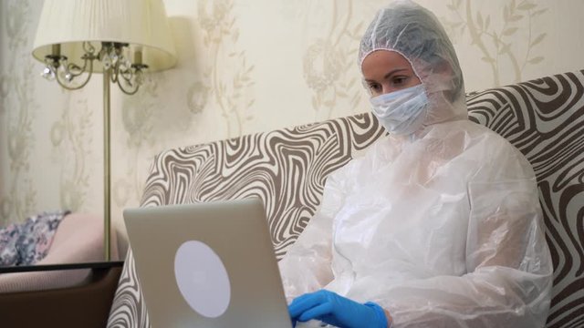 Medic nurse sitting on sofa in white protective suit, medical face mask, blue rubber gloves at home close up. Man is typing text, remote works, using laptop in living room during pandemic by lockdown