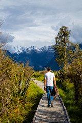 White man walking in a wooden path with mount Tasman and Mount Cook in the background in the surroundings of Lake Matheson, South Island, New Zealand