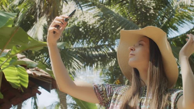 Cheerful lady making selfie photo on mobile phone over green palm tree background. Portrait of happy female tourist shooting herself outdoors in tropical nature slow motion. Smart technologies app