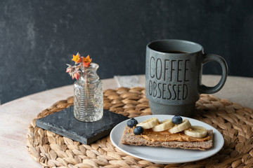 Peanut butter with blueberry, banana and sesame seeds on wholegrain rye crisp bread with mug of hot black coffee on a wooden table