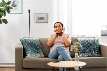 people, technology and leisure concept - happy young african american woman in glasses with headphones and smartphone sitting on sofa and listening to music at home