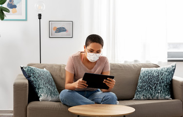 people, technology and leisure concept - young african american woman in glasses and face protective medical mask for protection from virus disease sitting on sofa with tablet pc computer at home