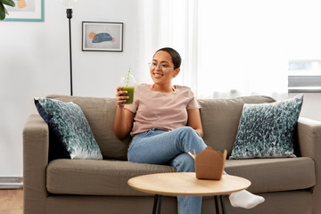 people, eating and leisure concept - happy smiling african american young woman sitting on sofa and drinking smoothie from plastic cup with paper straw at home