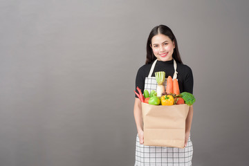 Fototapeta na wymiar Portrait of beautiful young woman with vegetables in grocery bag in studio grey background