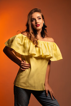 Studio portrait of young beautiful woman with long curly hair and perfect make up wearing a yellow blouse and red earrings on orange background. Text space.