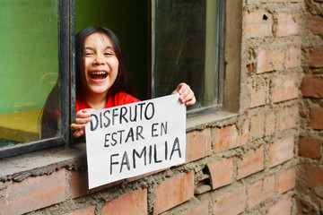 Naklejka na ściany i meble Little latin girl standing near the window and holding a sheet of paper that says in spanish 