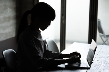 暗い部屋の中でパソコン作業をする若い女性のシルエット姿