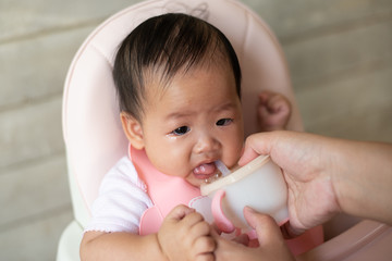 Little newborn baby sitting in baby chair  crying and refuses to drink water while mother tries to feed with a water cup.