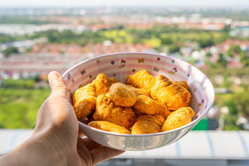 The hand that is holding the yellow fried nugget bowl Behind is a terrace view of the city and nature.