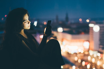 Woman is sitting near the window and using mobile phone at hotel room in the dark at the night.