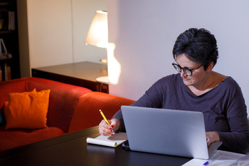 Signora con i capelli scuri e maglia colorata lavora in modalità smart working da casa sua con il portatile e prende appunti