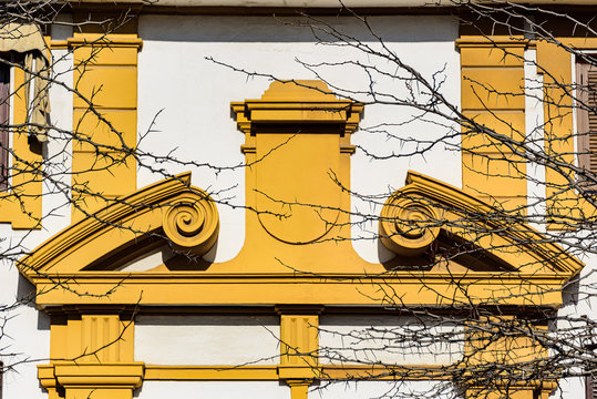 Yellow pediment over a doorway in curved and broken-apex style. The opening in the middle is facial-like molding which resembles a person flexing his arm muscles.
