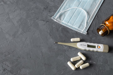 On the rough gray surface are a medical mask, digital termometr and a bottle of brown glass with pills poured out of it. COVID-19 concept. Horizontal orientation, selective focus, copy space.