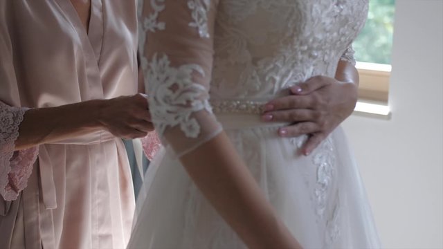 Mother in pastel morning robe helps her daughter put on wedding dress, slowmo