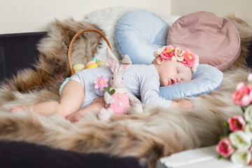 Little baby girl in flower crown, sleeping on a sofa on blue pillow and artificial fur, surrounded by easter decorations - bunny, tulips and basket with eggs