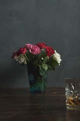 Vase with roses and a glass of whiskey on a wooden table.
