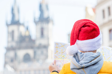 Naklejka premium Young boy wearing red santa's hat stands on the old town square in Prague and holds map. Empty space for text