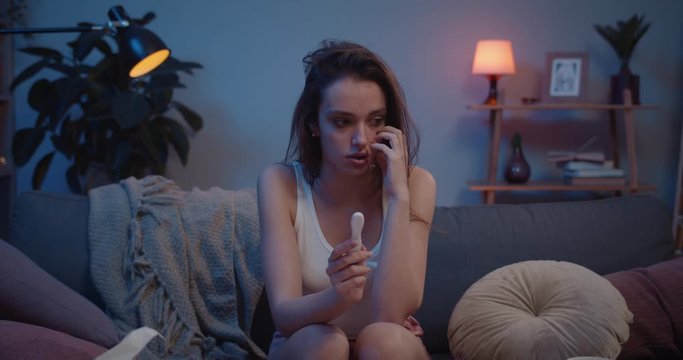 Front view of shocked sad girl finding out about having baby. Young unhappy woman sitting on sofa and worrying about unwanted pregnancy. Concept of fear and unplaned pregnancy