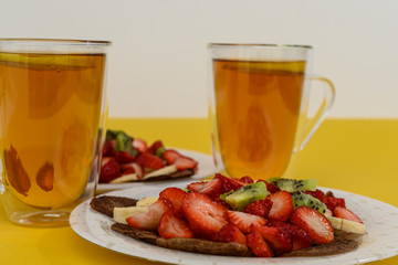 Fresh pancakes with chopped fruits, strawberries, bananas, kiwi and honey on light plates. Serving for two. Two cups of black tea with lemon. Healthy food, gluten free. Copy space. Yellow background
