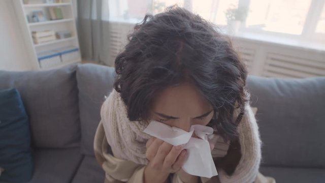 Close up of curly-haired Caucasian woman wearing warm scarf sitting at home, sneezing and coughing