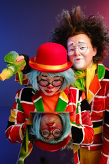 two clowns a man and a woman with bright makeup in colored costumes say they look at their reflection in the mirror. 