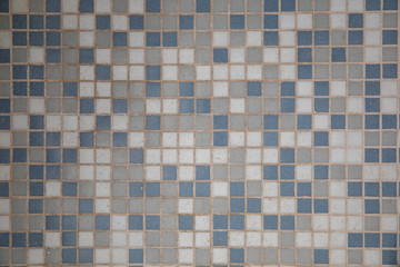 Close-up on a mosaic made of small gray, blue and white ceramic squares. It forms the floor of a lighted room with it is worn out due to numerous passages of people walking on it