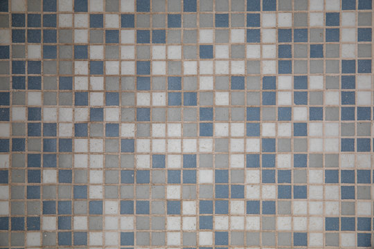 Close-up on a mosaic made of small gray, blue and white ceramic squares. It forms the floor of a lighted room with it is worn out due to numerous passages of people walking on it
