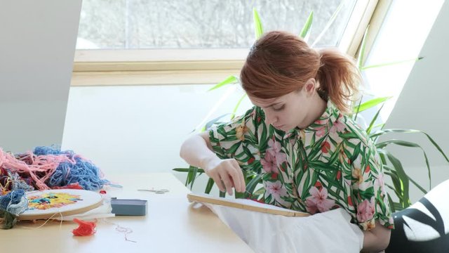 young pretty girl in flowered summer shirt sitting at table sewing with needle. embroidery hoop, fabric with colorful pattern. Traditional hobby, lifestyle. Needlework, handicraft. quarantine leisure