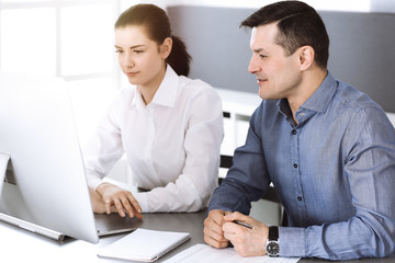 Fototapeta premium Cheerful smiling businessman and woman working with computer in modern office. Headshot at meeting or workplace. Teamwork, partnership and business concept