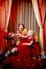 Family during a stylized theatrical circus photo shoot in a beautiful red location. Models mother and daughter posing on stage with curtain