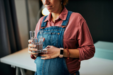 Side view of cute pregnant Caucasian woman standing in home office and drinking fresh water.