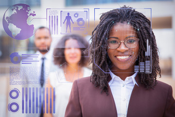 Cheerful young African American businesswoman with face detection infographics. Portrait of confident female boss smiling at camera while standing with multiethnic colleagues. Leadership concept