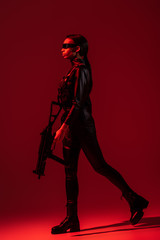 futuristic african american woman in glasses walking with assault rifle on red background © LIGHTFIELD STUDIOS
