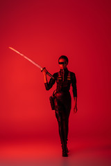 futuristic african american woman in glasses walking with sword on red background
