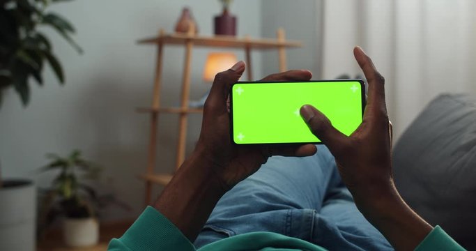 Man holding green screen smartphone in horizontal landscape mode with trecking markers while lyying on sofa . African guy watching video while looking at mock up screen.