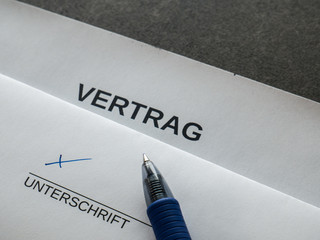 Symbolic pen and sheet of paper on a desk showing the text CONTRACT in German, below there is a...