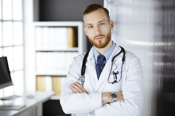 Red-bearded doctor standing straight in clinic near his working place. Portrait of physician....