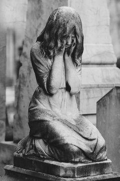 black and white sculpture of a angel girl in an old cemetery. Closeup of stoned angel with closed eyes and cross monument at cemetery. Graveyard old weathered stone sad baby statue on funeral.