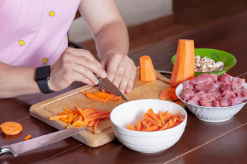Man hands cutting vegetables for cooking. Carrot and mushrooms, row meat, ingredients for stew or ragout. Classical brown wooden kitchen interior, pink shirt, black watch, modern male routine. 